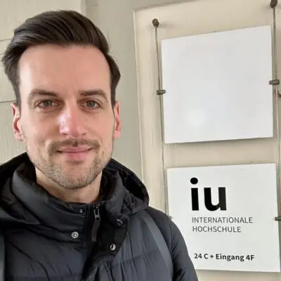 Dozent Thorben macht ein Selfie vor IU Logo am Campus